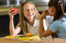 Mädchen mit Apfel in der Hand unterhält sich mit anderem Mädchen am Essenstisch. © Getty Images
